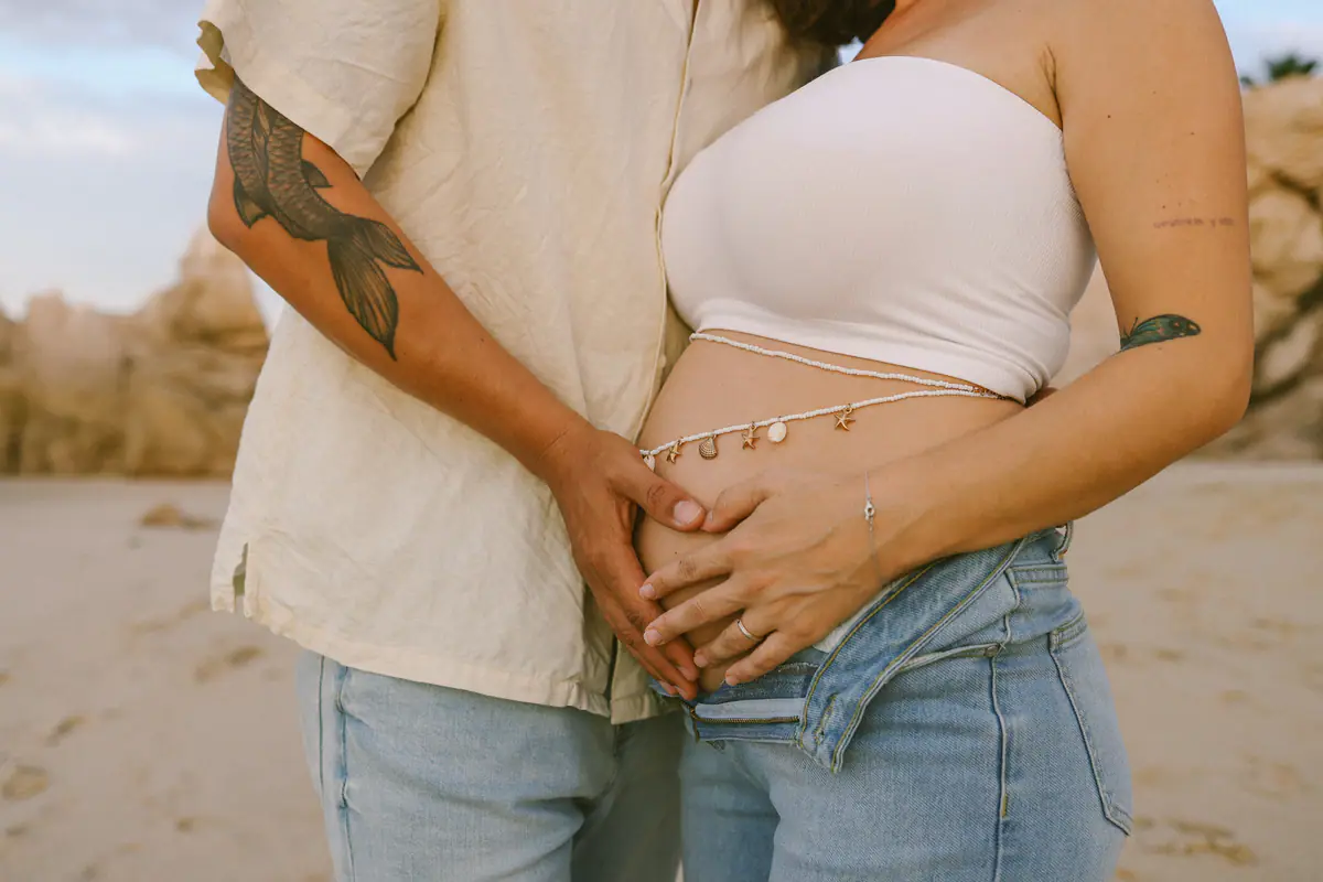 Two pairs of hands resting on her pregnant belly, close up on the beach