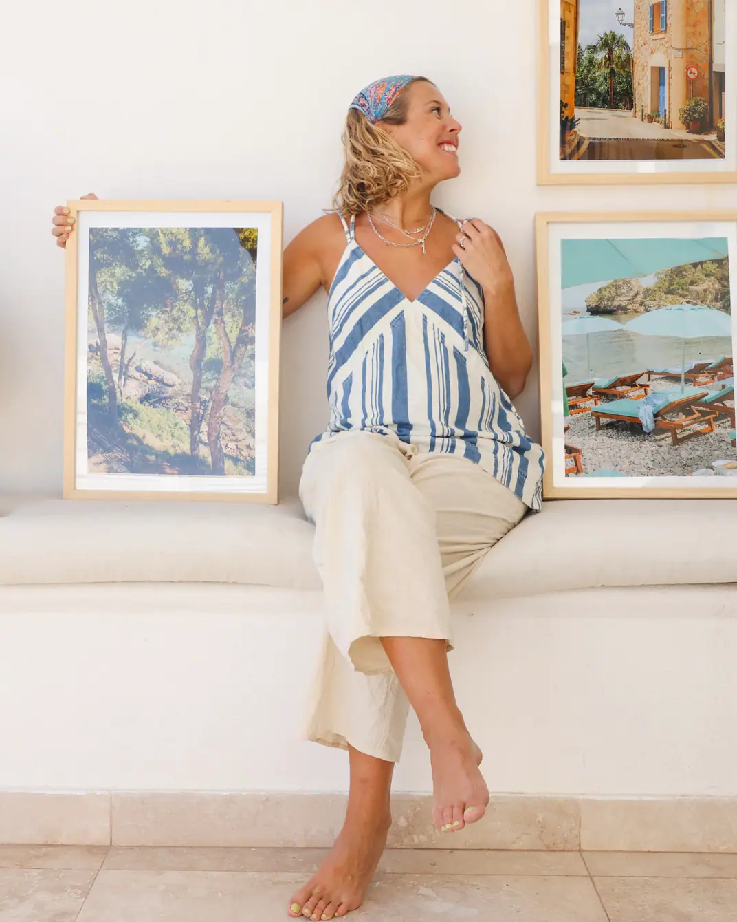 Carol Kuchman laughing, sitting barefoot on a white bench next to framed prints