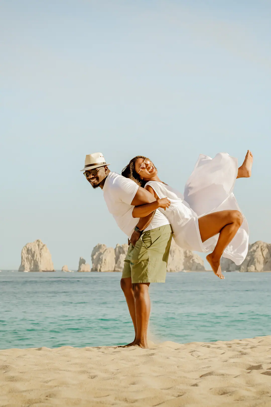 A couple laughing on the beach, she jumps into his arms with El Arco and the sea behind them