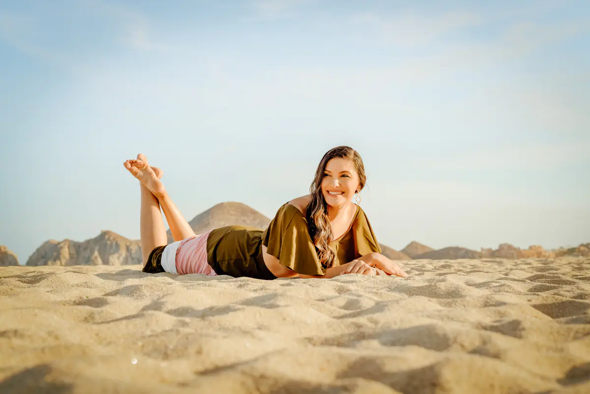 A woman lying on the sand smiling, with the Land's End rocks behind her